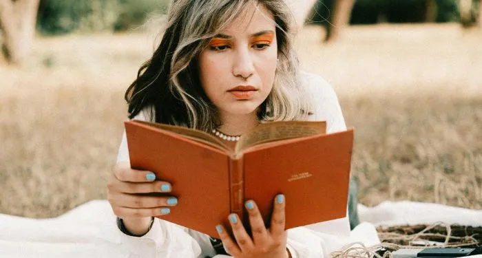 fair-skinned woman reading an orange book while lying on a blanket in the grass